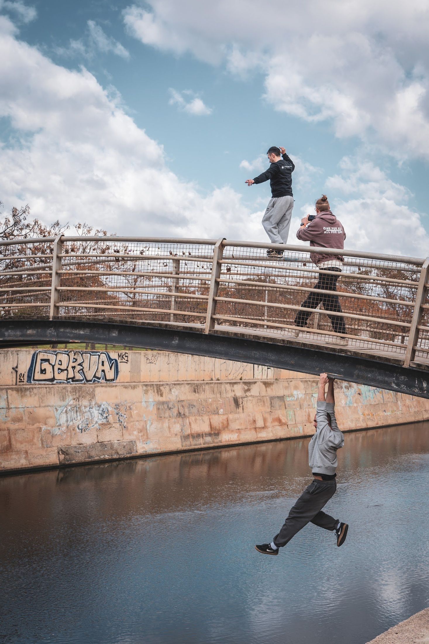 One man balances on the railing of a footbridge while another hangs from its underside above a canal.