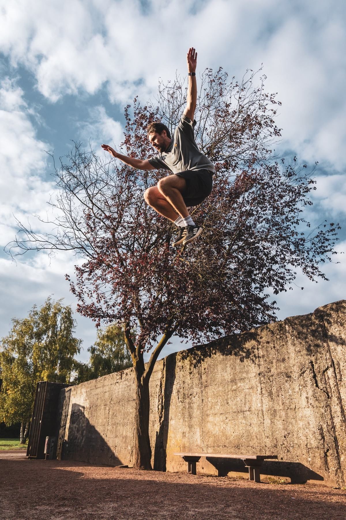 A man jumps high off a concrete wall, arms spread, in front of a red-leaved tree and a cloudy sky.
