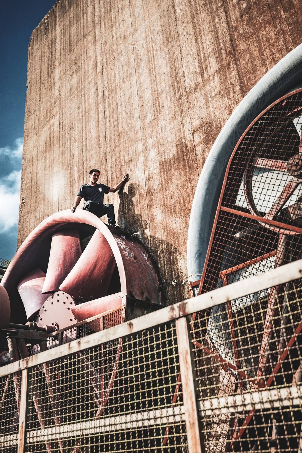 Minh crouches on top of a giant rusty industrial fan against a weathered red wall.