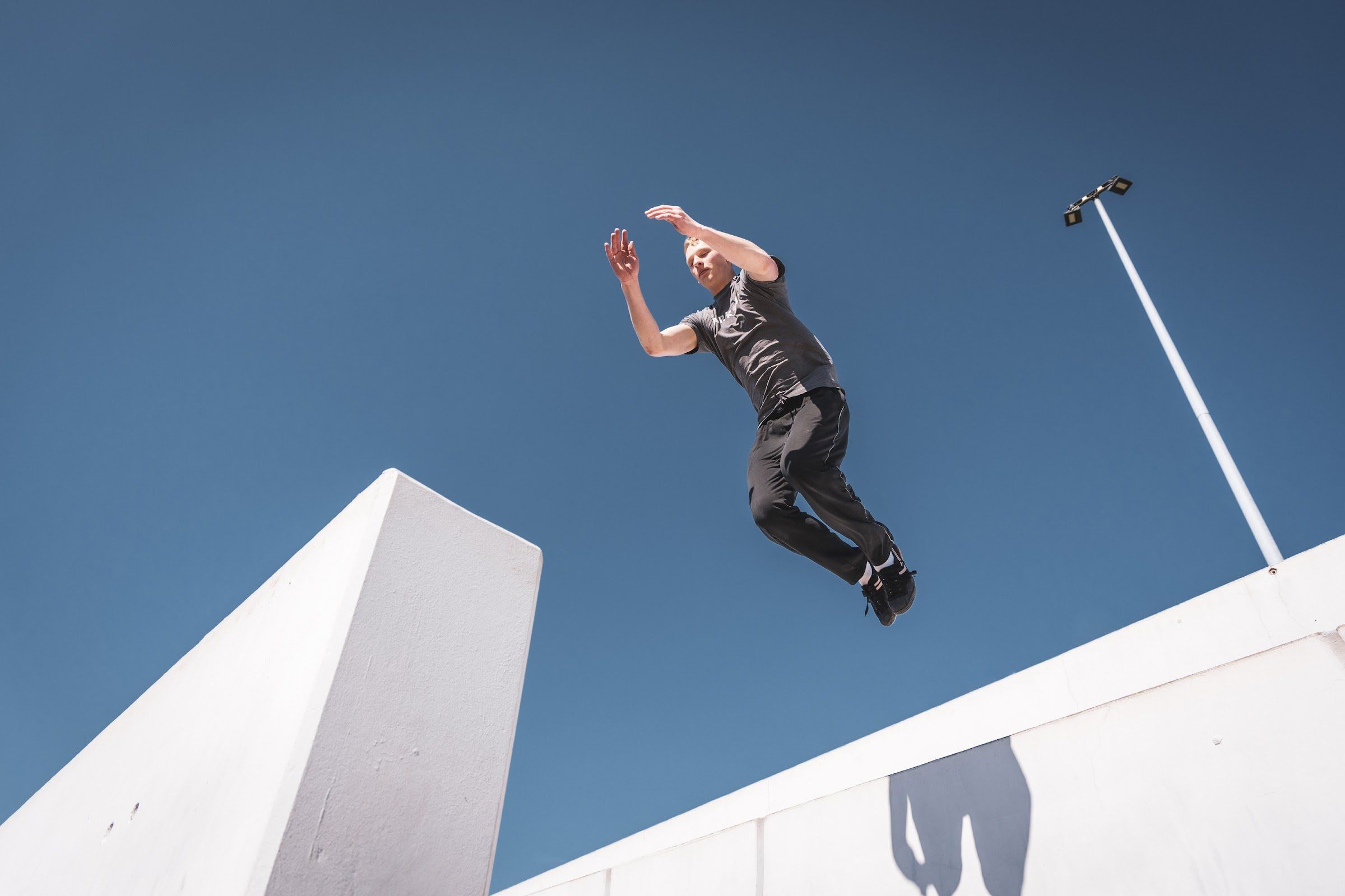 A man in a grey t-shirt leaps between two white concrete walls against a clear blue sky.