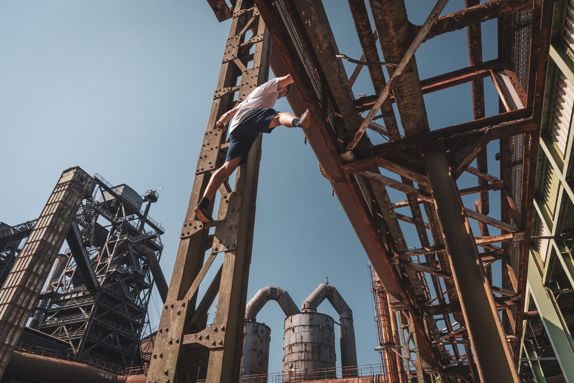 A man climbs a rusty steel girder high above, with blue sky and an old blast furnace behind.