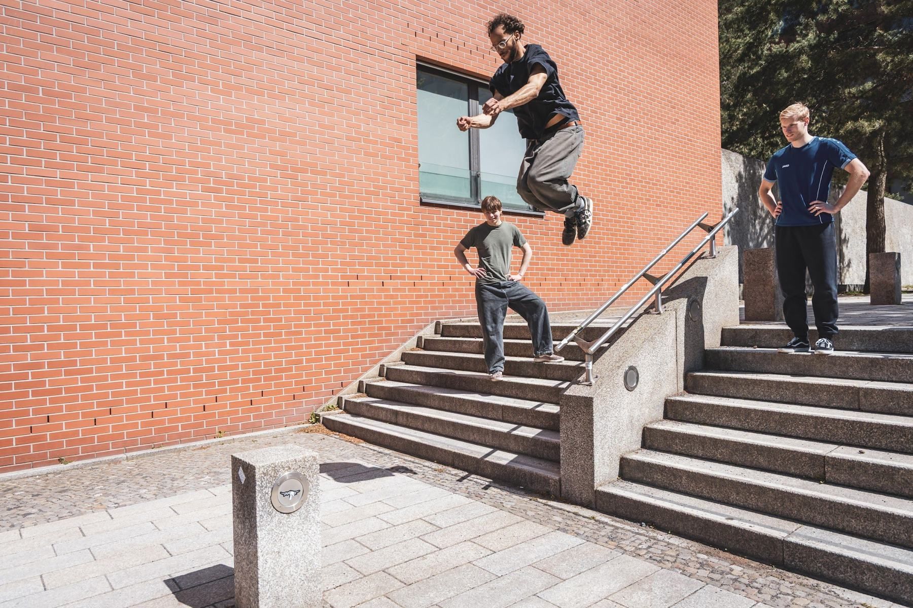 An athlete jumps over a stone staircase in front of a red brick wall while two people watch.