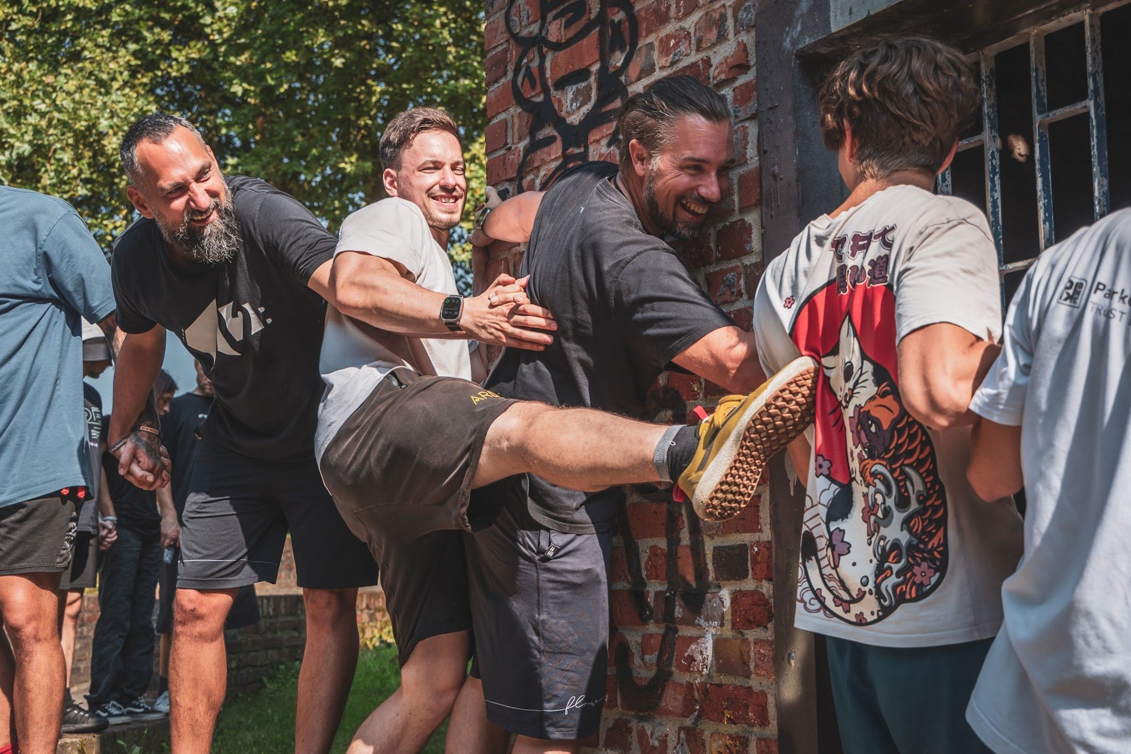Laughing coaches link arms in a game against a sunny brick wall, one kicking his leg high.