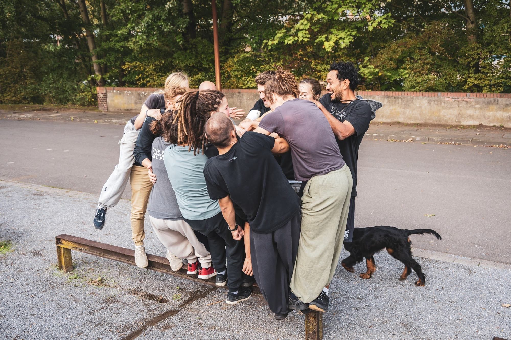 A tight cluster of people hold on to each other to fit on one low metal rail while a black dog walks by.