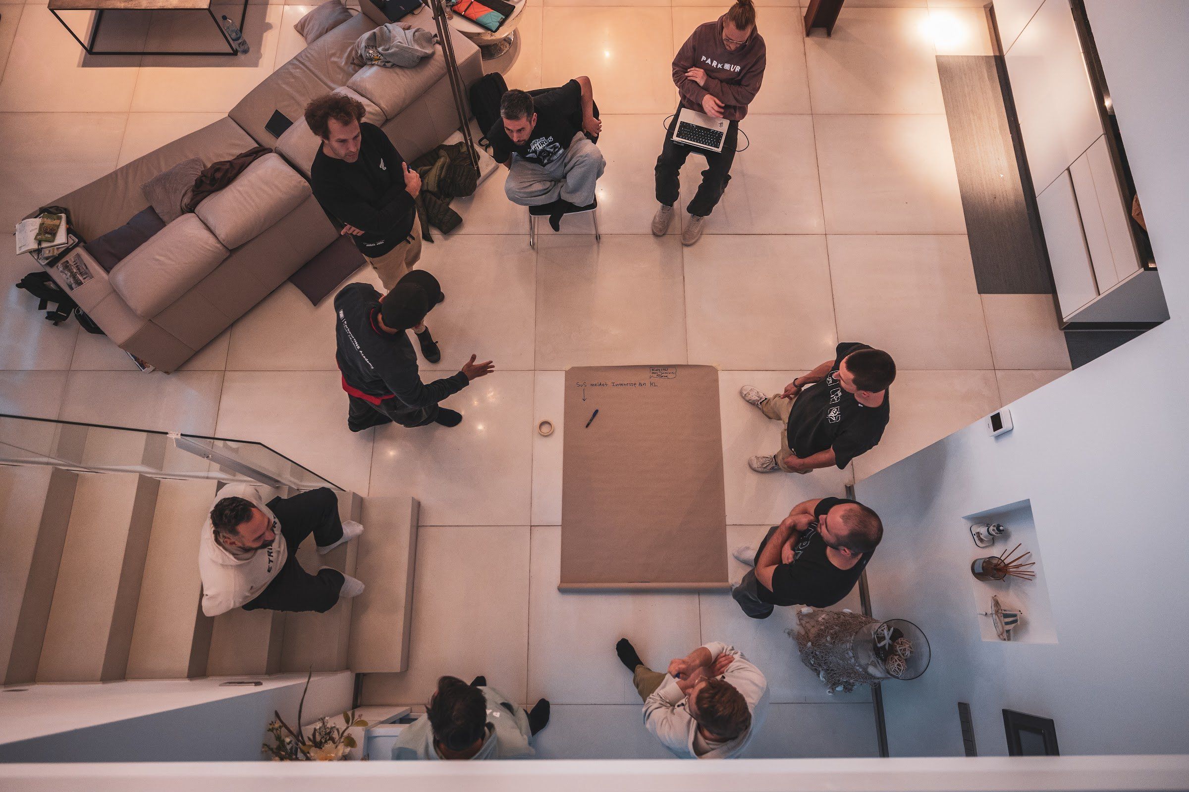 Seen from above, a group gathers around a large sheet of paper on the floor of a warmly lit room.