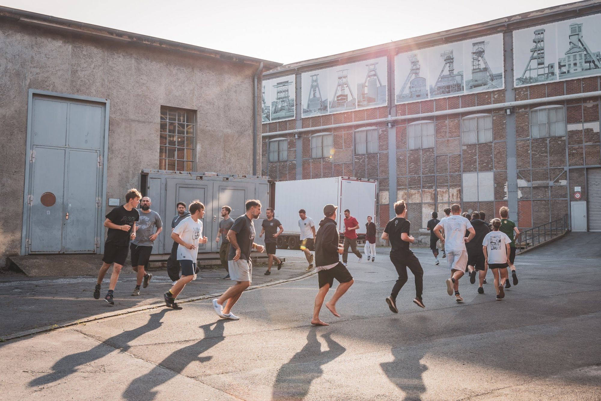 A large group runs across a sunlit industrial courtyard, casting long shadows.