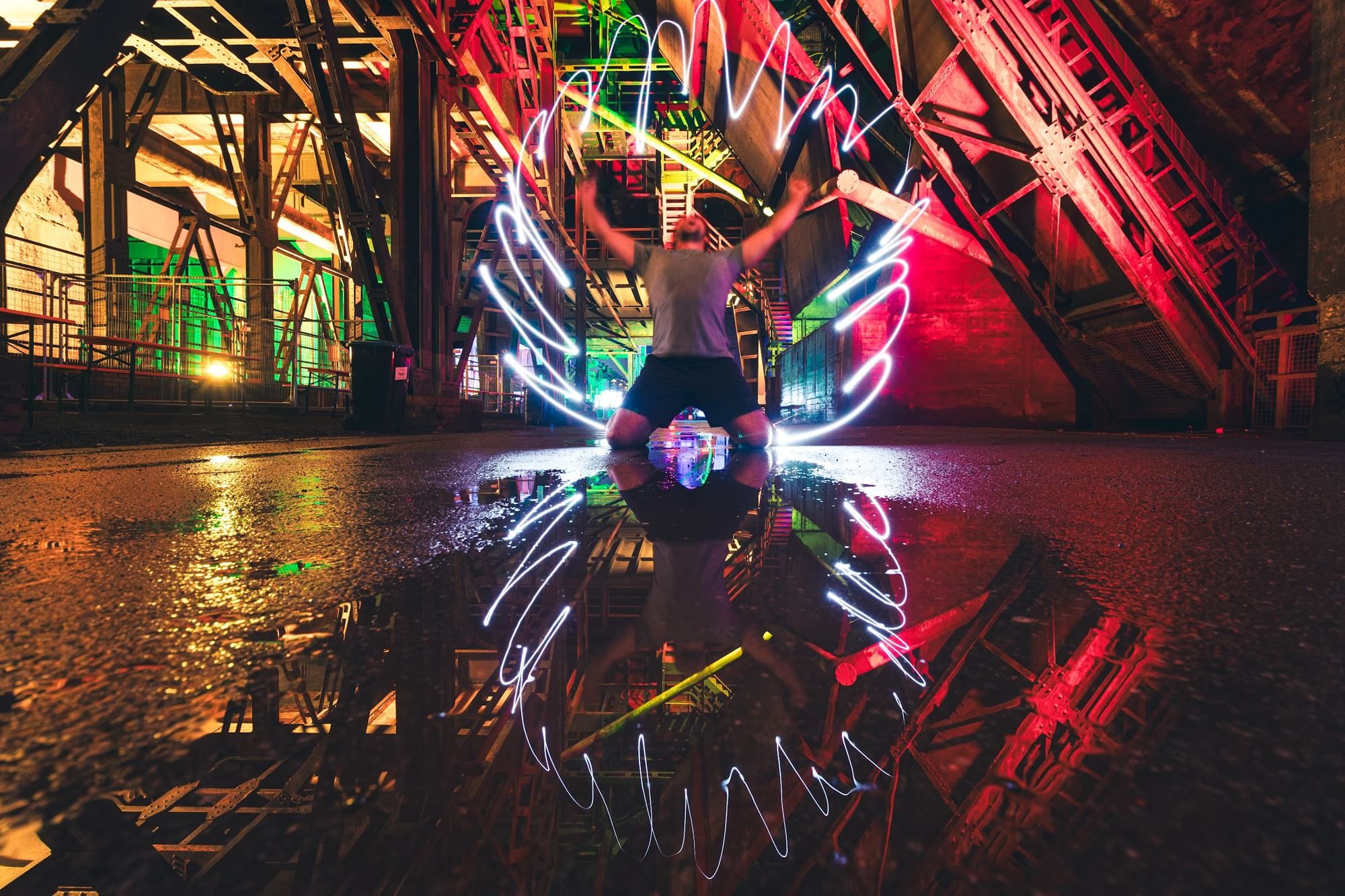 A man kneels in a puddle at night, framed by glowing light-painted wings and coloured industrial lights.