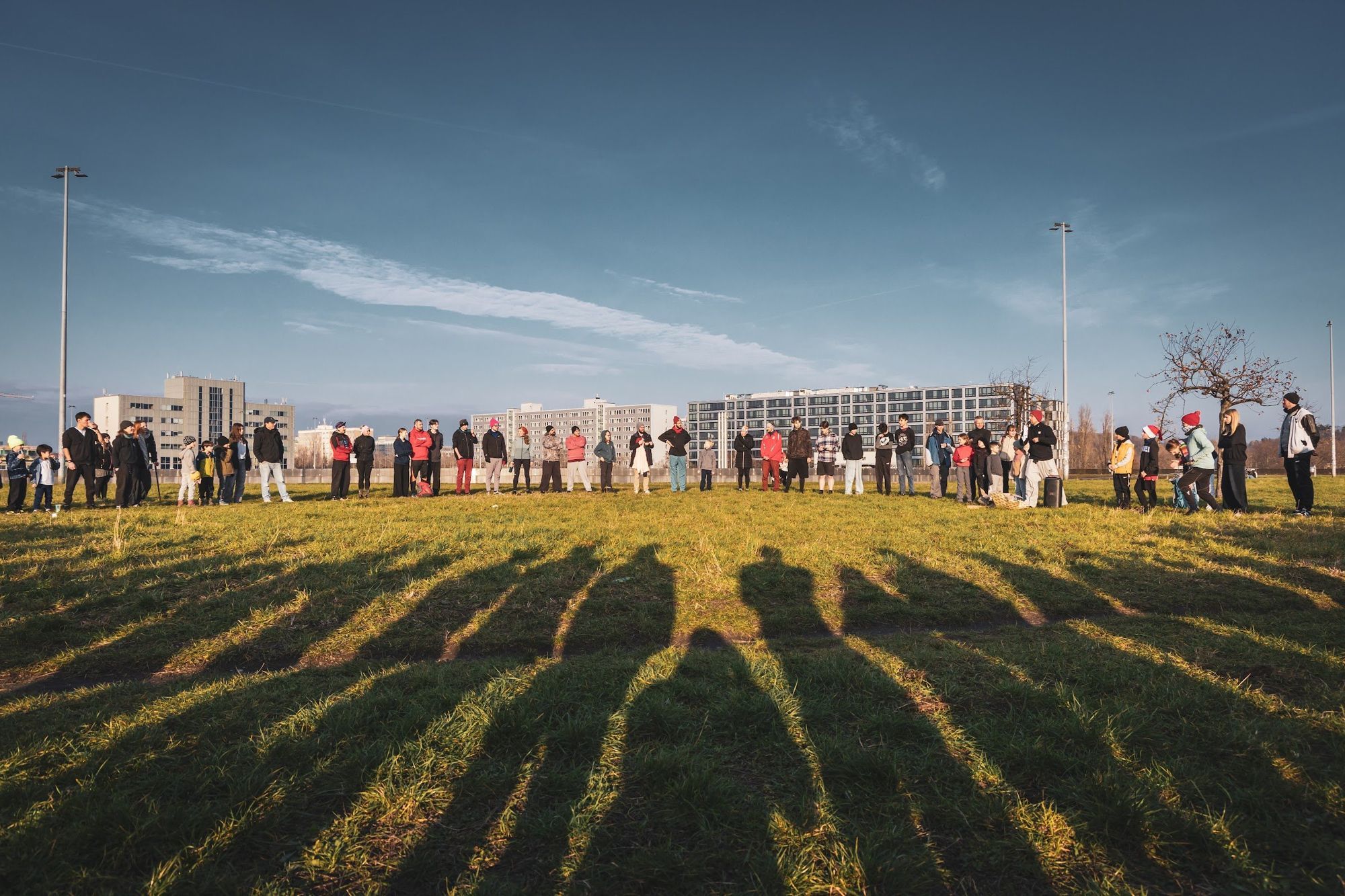 A large group stands in a circle on a sunny field, their long shadows reaching across the grass.