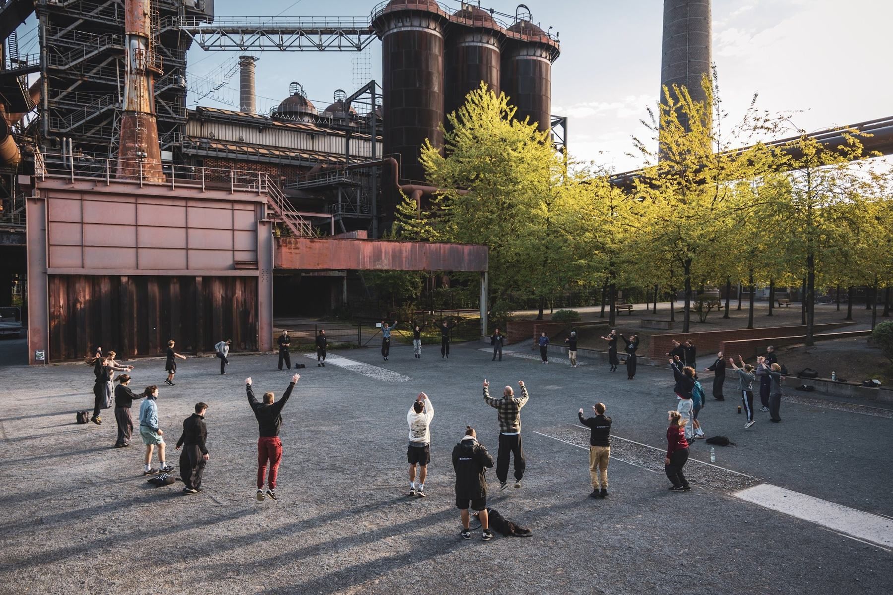 A large group warms up in a circle in front of rusty blast furnace towers, several people jumping with arms raised.