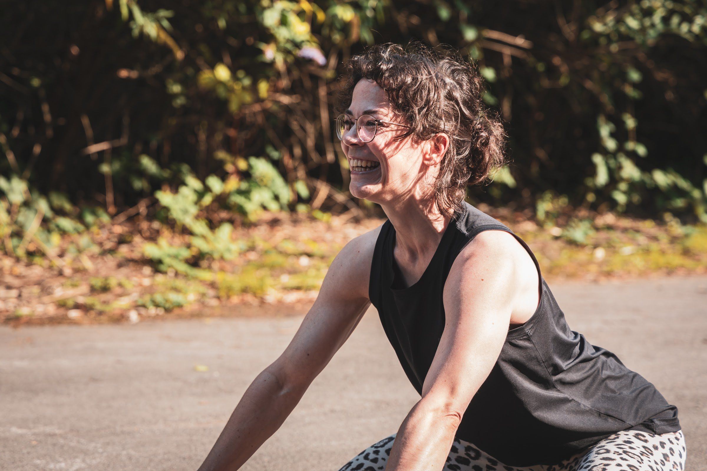 A woman with curly hair and glasses laughs during a stretching session in warm sunlight.