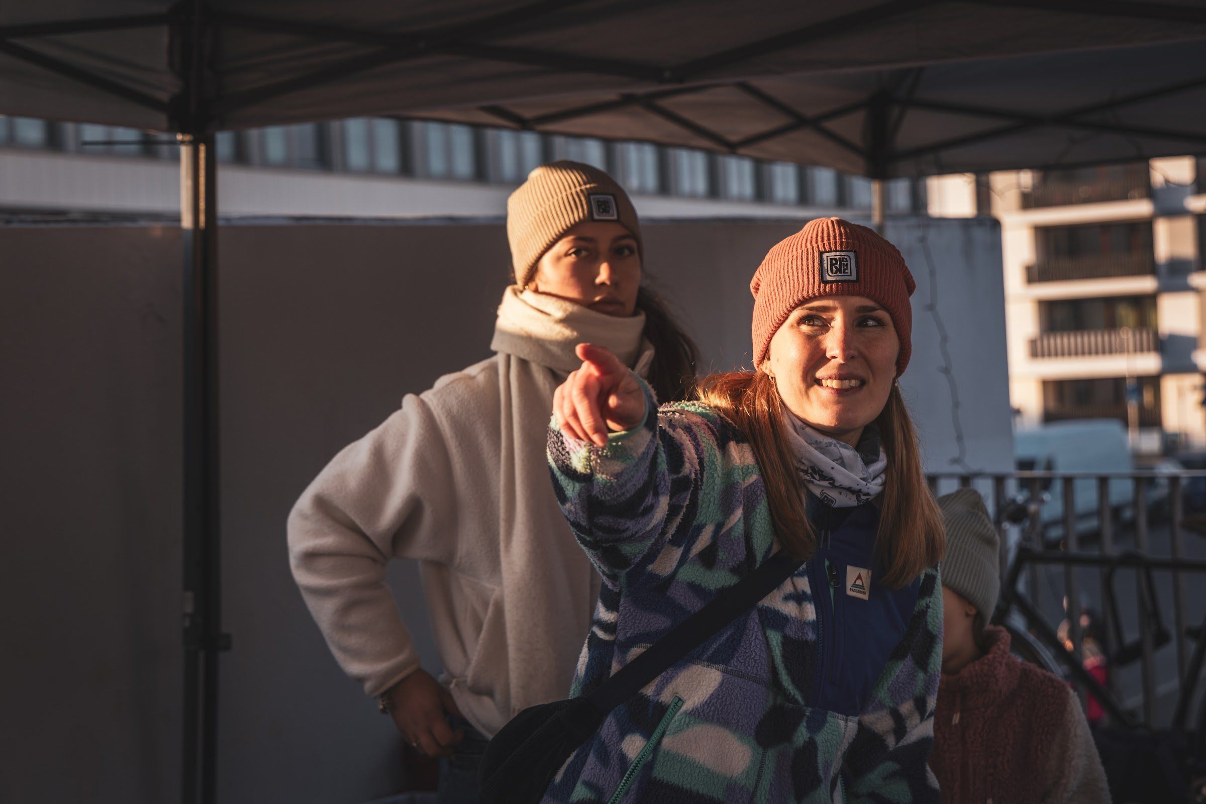 A smiling woman in a rust beanie points past the camera in low winter sun.