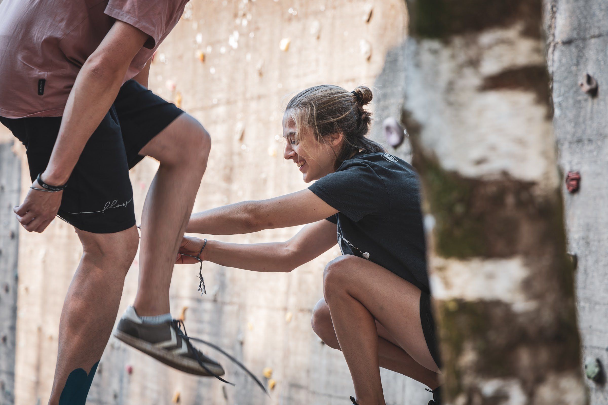 A smiling coach steadies a climber's leg at a concrete climbing wall.