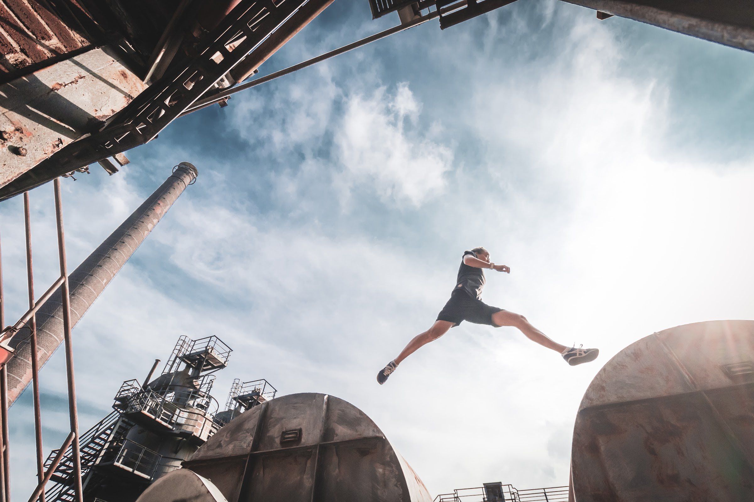 A woman leaps in a wide stride across the sky between two rusted industrial tanks.