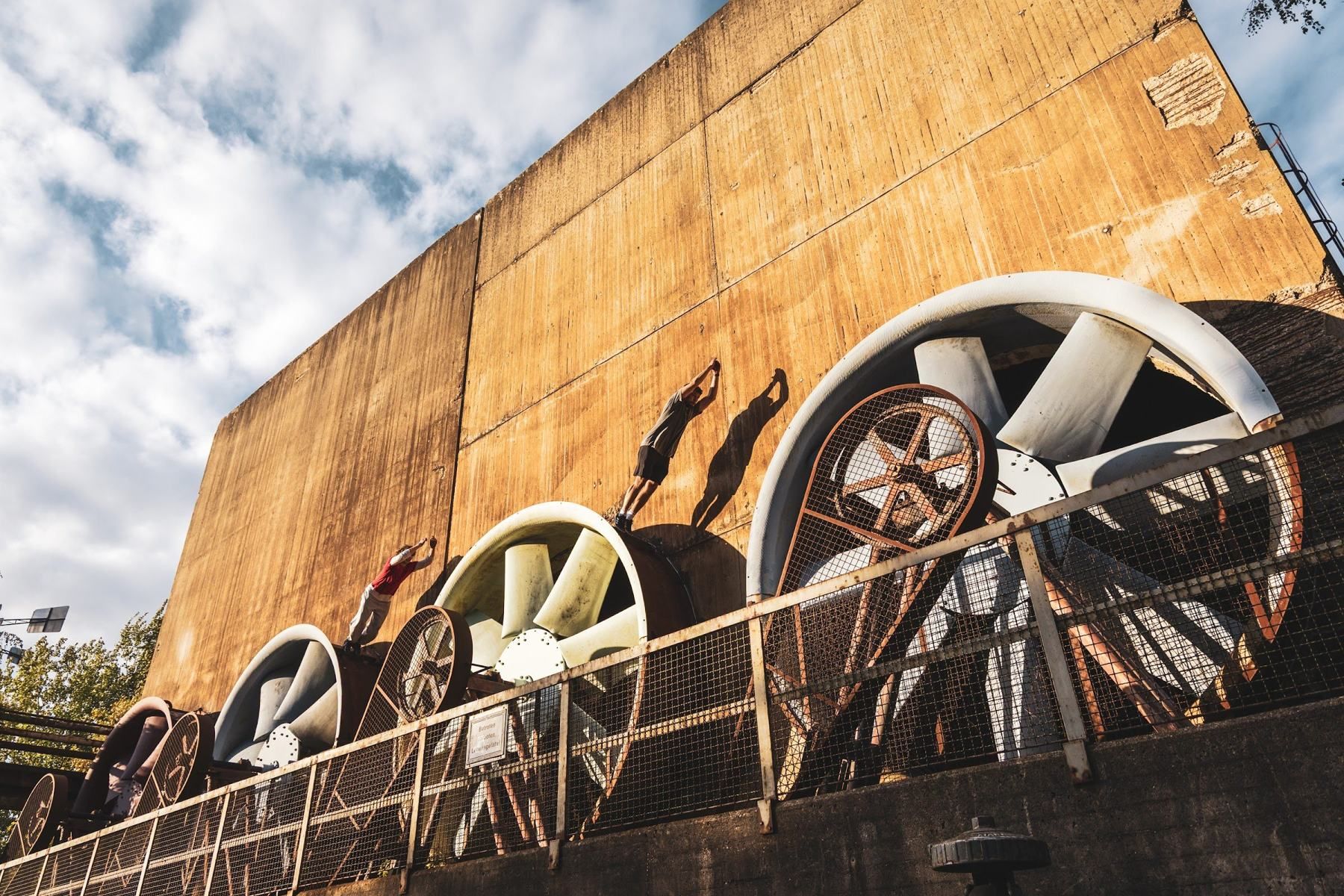 Two people climb onto giant industrial fans in front of a towering rust-coloured wall.